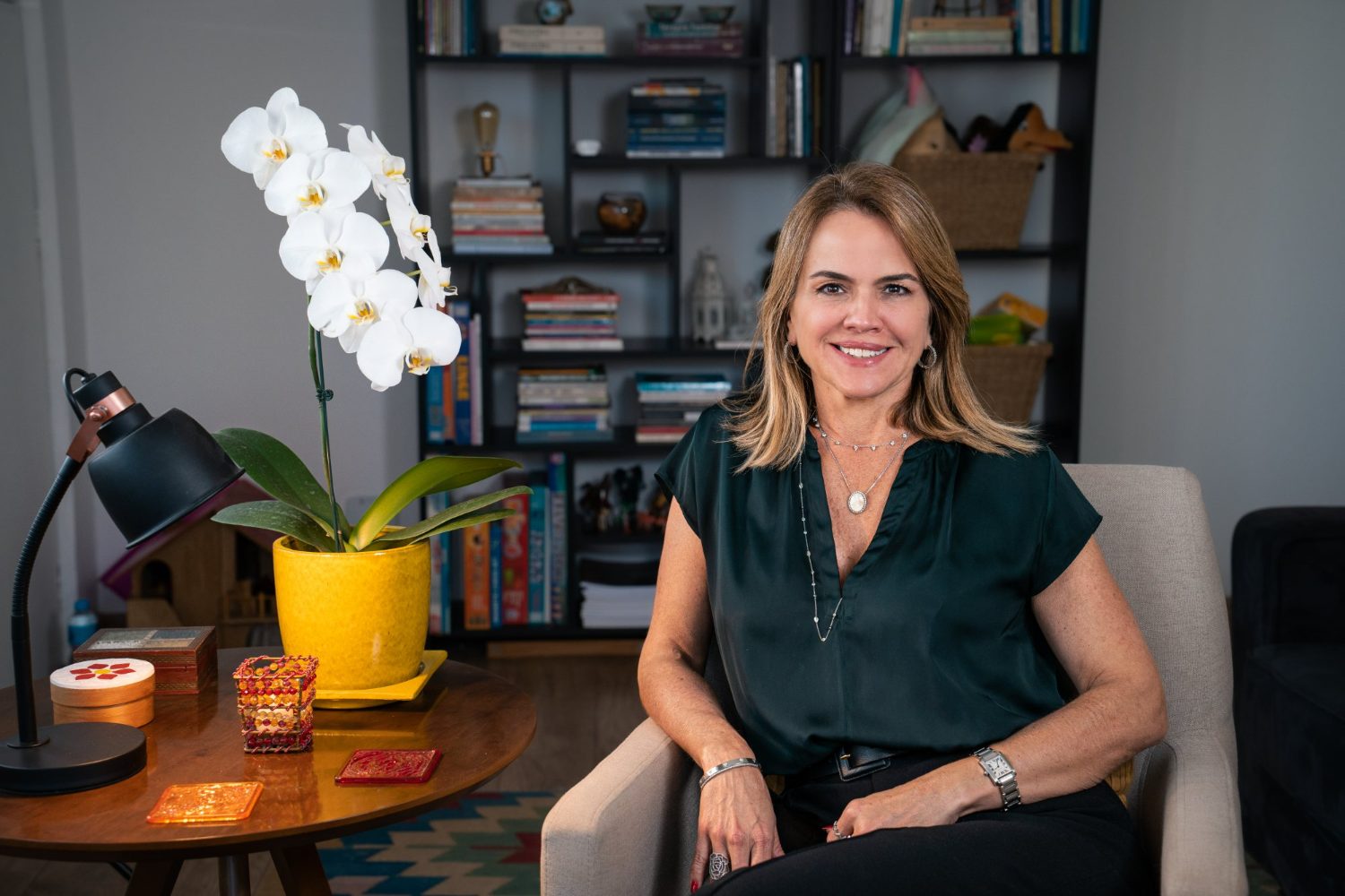 Consultório com poltrona, estante de livros, luminária e uma orquídea branca, com Lecticia Raposo sentada ao centro